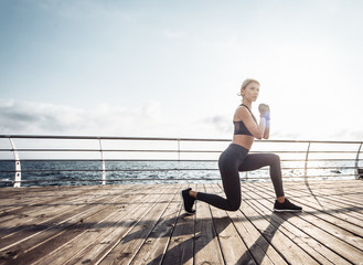 Fototapeta na wymiar Healthy woman training on seaside promenade. Young attractive woman in sportswear does lunges exercise on the beach at sunrise. Warm up
