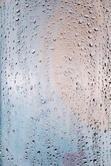 Drops of water flow down the surface of a transparent glass on an abstract blurred bright colored background.