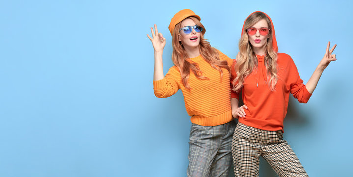 Two Lovable Embracing Fashionable Woman Sisters In Trendy Orange Yellow Outfit. Carefree Beautiful Funny Stylish Friends Smiling Shows Peace Sign On Blue. Happy Fashion Girl, Colorful Positive Banner