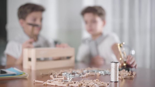 Blurred Caucasian Children Sitting At The Background, One Boy Hammering Nail, His Twin Brother Moving Away Sandglass. Woodworking, Carpentry For Children, Hobby. Focused On Foreground.