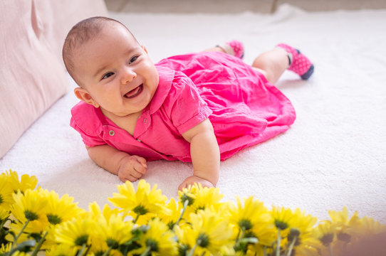 Baby in pink dress on white texture, hands resting next to yellow flowers laughs funny and shows tongue