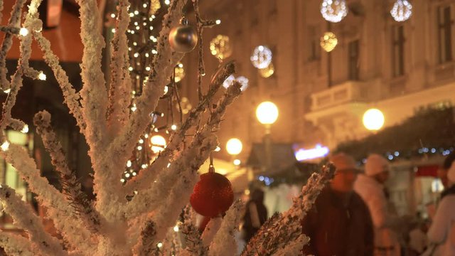 CLOSE UP DOF: Beautiful white Christmas tree stands near a busy street in Zagreb. Tourists explore the picturesque advent market in Europe. Idyllic shot of red ornaments hanging from snowy spruce tree