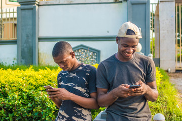 two young black men using their mobile phones outdoor, texting, and browsing the internet, smiling while using their phones