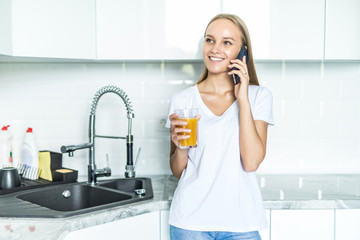Young joyful woman drinking orange juice while talking mobile phone and standing near a kitchen table
