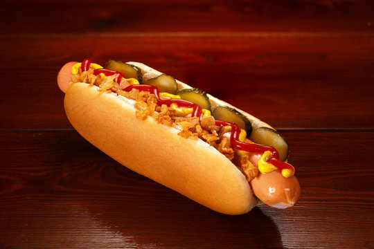 Danish hot dog with pickled cucumbers, fried onions, ketchup and mustard. On a wooden table.