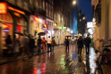 Blurry motion image of people walking on Warmoesstraat street in Amsterdam. It is one of the main shopping streets with cafes, restaurants and shops. It is a rainy summer night. Youth culture concept