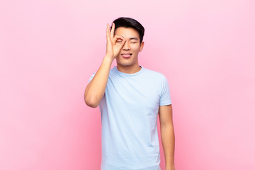 young chinese man smiling happily with funny face, joking and looking through peephole, spying on...