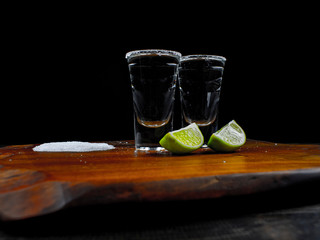 Two tequila silver shots with fresh lime and sea salt on wooden board and dark background