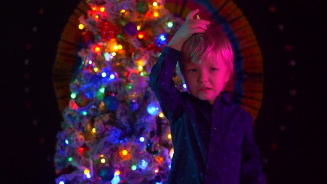 funny blond kid in bright stockings stands in front of Christmas tree with candle. Bright lights of garlands blink. Great mood before the New Year. 2020. Christmas Eve. Mixed media. Shallow focus