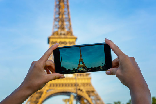 Eiffel tower in Paris under sunlight and blue sky. Famous popular tourist place in the world. Tourist in Paris visiting landmark sightseeing in France, woman taking photo on mobile phone.