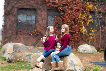 Two young teenager girls sitting on the rock, drinking coffee. Sisters, friends happy smiling, laughing, talking. Fall autumn time.