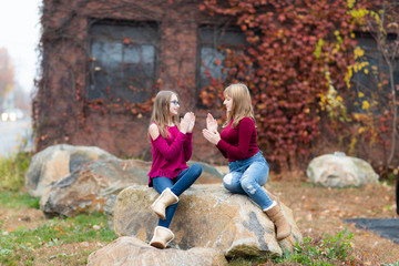 Obraz na płótnie Canvas Two young teenager girls sitting on the rock, drinking coffee. Sisters, friends happy smiling, laughing, talking. Fall autumn time.Playing clapping hands 