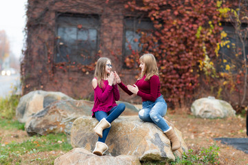 Two young teenager girls sitting on the rock. Sisters, friends happy smiling, laughing, talking. Fall autumn time. Playing clapping hands 