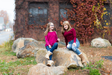 Two young teenager girls sitting on the rock, talking. Sisters, friends happy smiling, laughing, talking. Fall autumn time.