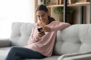 Fototapeta premium Smiling girl sit on sofa receiving pleasant message on smartphone