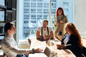 A group of colleagues having coffee, 2 men and 2 women chatting. Airy and spacious office with panoramic windows, filled with sunlight. Employees with coffee cups dressed in smart casual outfits.
