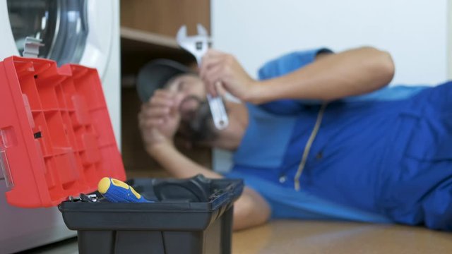 Handyman in the kitchen. The plumber lies on the floor under the kitchen faucet and takes his tools from the toolbox.