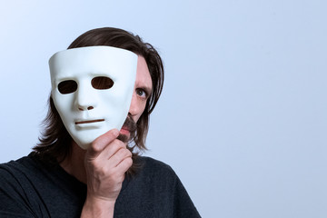 A long-haired man with a mustache holds a white mask near his face and looks out with one eye. The...