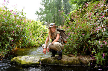 A young girl dresses up as an explorer. She wears a safari hat and wears khaki clothing. She is seen in a  river environment.