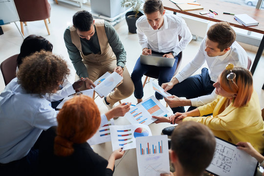 Diverse young people of business team gathered to discuss business startup in modern office, sit together in circle holding papers with diagrams. active discussing