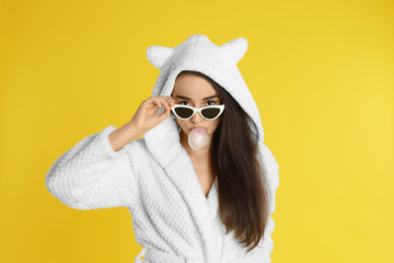 Young woman in bathrobe and sunglasses blowing chewing gum on yellow background