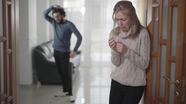 Blurred Caucasian man yelling at his girlfriend or wife from background. Depressed woman holding pregnancy test and crying. Unwanted pregnancy, relationship problems.