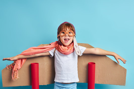 Cheerful Smiling Kid Boy Imagine Himself As Pilot, Professional Of Aviation. Wearing Cardboard Plane On Back And Pilot Helmet. Isolated Over Blue Background