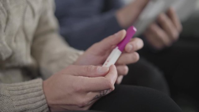 Close-up side view of female Caucasian hands holding affirmative pregnancy test. Blurred male hands at the background using tablet. Wife deciding how to tell husband about pregnancy.