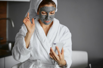 beautiful caucasian woman in white bathrobe and towel use beauty procedures sitting at home after shower with smartphone