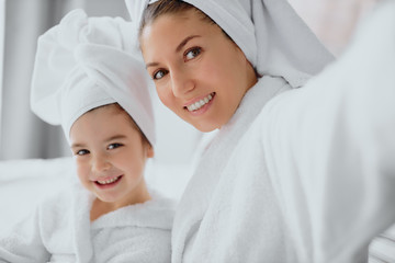 portrait of beautiful female wearing white bathrobe and towel sit looking at camera and smile, bedroom background