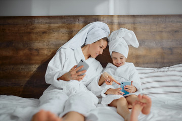 cute young mother and daughter spend time with each other using smartphone, wearing bathrobe and towel
