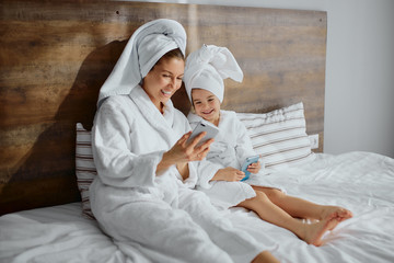 young modern beautiful woman wearing bathrobe and towel lie on bed with daughter using smartphone