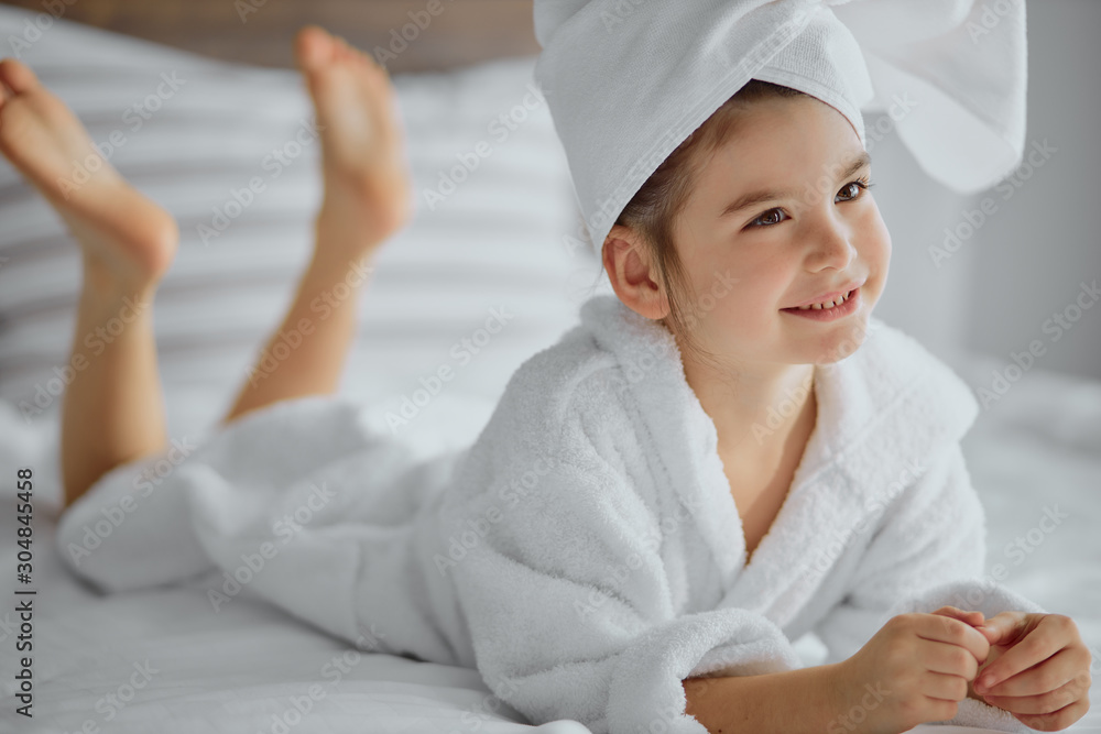 Wall mural awesome cute little girl of caucasian appearance wearing bathrobe and towel, lies on bed