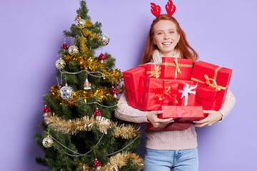 Pretty attractive girl carrying various Christmas gifts and presents in hands, looking straight with charming smile, expressing happiness, satisfied with preparation to New Year's Day