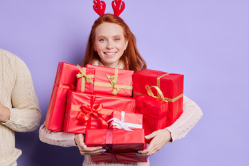 Smiling playful girl happily carrying many beautiful Christmas present, finding lots of presents under Christmas tree, wearing funny red horns, looking at camera with cheerful face, happy celebration