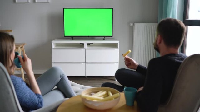 Man and woman are sitting in chairs, watching TV with a green screen, drink tea, eat snacks and talk. Back view. Chroma key. Timelapse