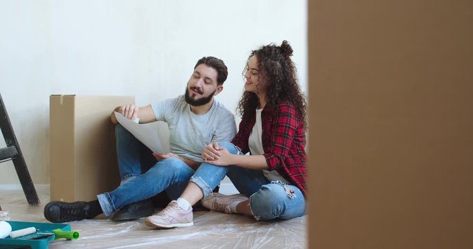 Just-married Caucasian couple sitting on the floor in the flat which being repaired and choosing the color for walls in the new room.