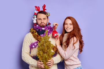 Portrait of tired boyfriend decorated with Christmas baubles and tinsel, happy woman playing with angry man, teasing and kissing, expressing happiness