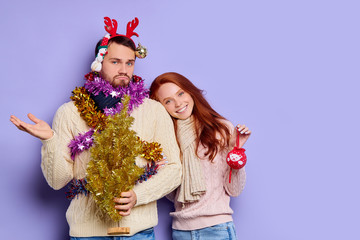 Upset doubt guy standing decorated with different Christmas baubles, spreading hands sideways with puzzled expression, redhead girl putting head on male shoulder with satisfied expression