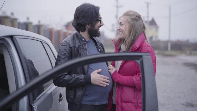 Smiling Caucasian blond woman caressing her husband's pregnant belly and imitating listening baby hitting. Cheerful couple exchanging their social roles. Joking, funny.