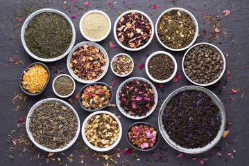 Assortment of dried tea leaves, fruit and herbs  in bowls on dark, stone background, top view