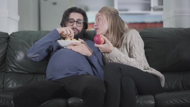 Funny caucasian couple sitting on couch and watching TV. Pregnant husband eating chips and his young beautiful wife biting apple. Family exchanging social roles. Joking, fun, kidding.