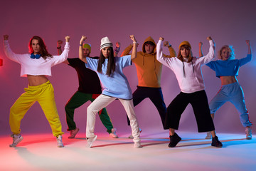 Fototapeta na wymiar Group of energetic street dancers in sportswear, standing on brightly lighted stage, raising hands up, keeping legs wide, performing dance movie, moving synchronically