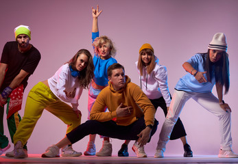Active group of modern style dancers demonstrating different gestures, young beard man in yellow cap showing tongue looking at camera, sport and urban culture concept