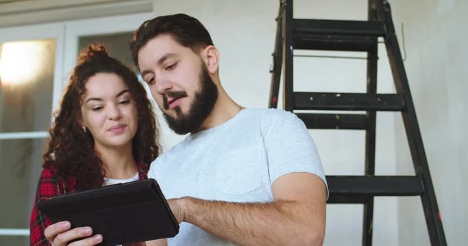 Young Caucasian good looking happy couple watching video or photos on the tablet computer while moving in the new apartment and doing renovation in it.