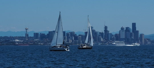 Two sailboats sailing in front of Seattle skyline and waterfront area including Elliott Bay as seen looking from Elliott Bay