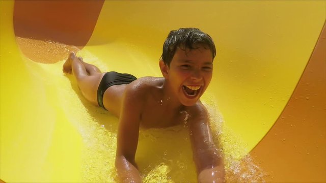 Funny laughing boy rides down orange slide on his stomach in a water park. Boy in a swimsuit laughs and slides into the water raising spray