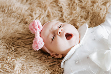 A three-month-old girl lies on a brown blanket wearing a white dress and a pink headband. Photo from above. The child yawns and looks at the camera