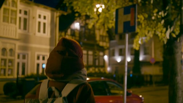Young Woman with Backpack Walks in Dark Street with City Lights in Late Evening or Night in Autumn or Winter. Tourist Lady in Red Jacket and Hat Walks Outside. Houses and Cars. 4K Backside Medium Shot