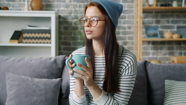 Slow motion of pretty girl in glasses and hat drinking holding mug at home relaxing sitting on sofa alone. Drinks, modern lifestyle and millennials concept.